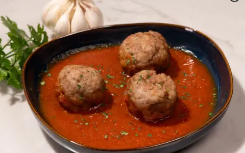 Tres albóndigas caseras en una rica salsa de tomate, adornadas con perejil fresco. Un plato reconfortante que evoca la cocina tradicional.