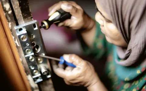 Une personne en hijab utilise un tournevis pour enlever vieux gonds d'une porte en bois.
