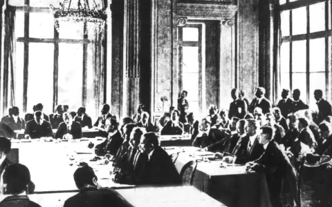 Männer in Anzügen sitzen an einem langen Tisch in einem prunkvollen Saal. Sie diskutieren die Bedingungen des Versailler Vertrags.