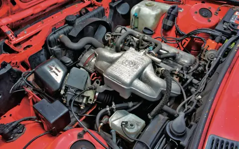 Silniki BMW E30 w czerwonej komorze silnika. Widoczny kolektor ssący z logo BMW, chłodnica, akumulator i inne elementy układu napędowego.