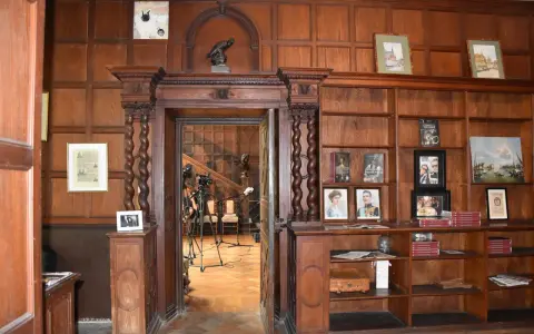 Wnętrze biblioteki w **zamku w Lubniewicach**. Drewniane boazerie, regały z książkami i portretami, a w przejściu widok na schody i sprzęt nagraniowy.