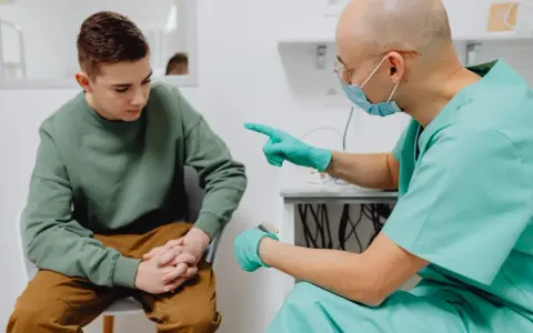 Lekarz w zielonym fartuchu i masce rozmawia z młodym pacjentem, wskazując palcem. Młody mężczyzna ma zmartwioną minę, być może z powodu obrzęku penisa.