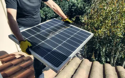 Person med gula handskar håller en solpanel på ett tegeltak, redo att koppla solceller till batteri för grön energi.
