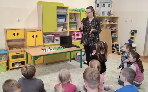 Nauczycielka prowadzi zajęcia kreatywne dla dzieci. Na stole przed nimi leżą klocki Lego, a dzieci z uwagą słuchają.