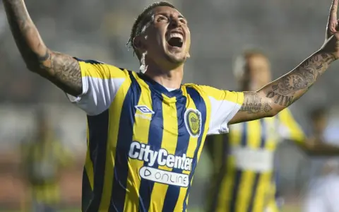 Jugador de Rosario Central celebra un gol, con los brazos en alto y la boca abierta. Sus tatuajes son visibles en sus brazos.
