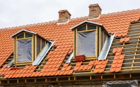 Trwa wymiana okna dachowego. Dach pokryty czerwona dachówką, widoczne drewniane elementy konstrukcji i nowe okna z folią.