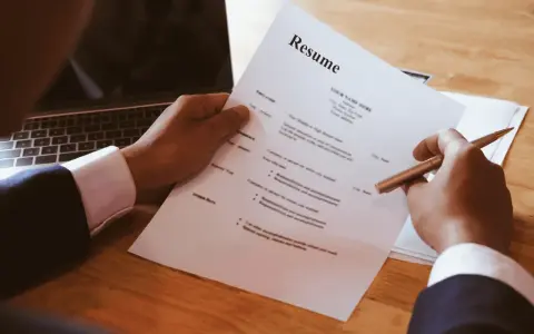 Dłoń trzymająca długopis nad dokumentem CV obok laptopa.