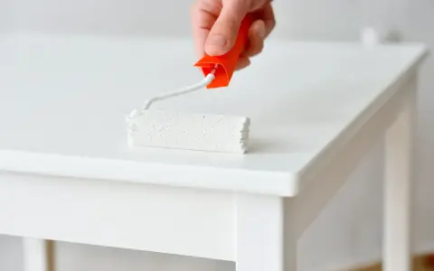 Main blanche d'un meuble laqué, un rouleau de peinture blanc est appliqué.
