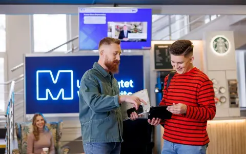 Dwóch studentów dyskutuje o kierunkach studiów WSB, trzymając laptopa i tablet. W tle widoczna jest neonowa litera "M" i ekran informacyjny.