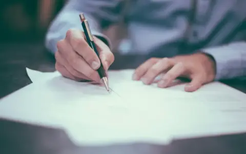 Dłoń pisze długopisem na dokumencie, przygotowując wpis do księgi wieczystej.