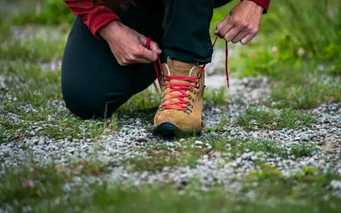 Osoba klęczy i sznuruje brązowy but trekkingowy z czerwonymi sznurówkami. To dobry przykład, jak zmierzyć stopę przed zakupem obuwia.