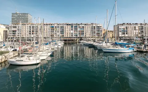 Barche a vela ormeggiate nel porto di Tolone, con edifici residenziali sullo sfondo e il cielo azzurro.