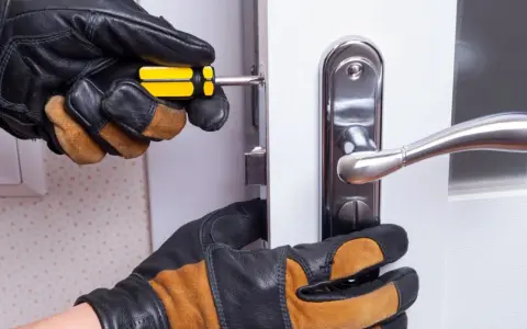 Mains gantées ajustent un loquet serrure bloqué sur une porte blanche avec un tournevis.