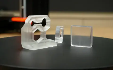 Three clear 3D printed objects, one complex bracket and two simpler shapes, sit on a black surface.