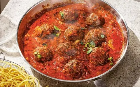 Frikadeller i tomatsås som puttrar i en panna, med en sidokorg med spaghetti.