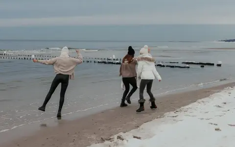 Trzy dziewczyny cieszą się zimowym dniem, gdzie zimą nad morze. Spacerują po plaży, na której leży śnieg, a fale delikatnie rozbijają się o brzeg.