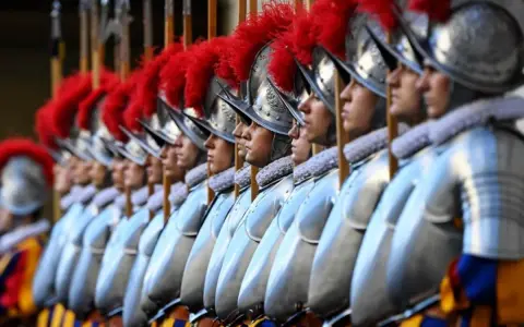 Guardie svizzere in fila, con elmi e piume rosse. Curiosità sulla loro uniforme storica e il loro ruolo.