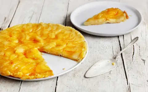 Eine goldbraune Tarte Tatin mit karamellisierten Äpfeln auf einem Teller, daneben ein Stück auf einem weiteren Teller und ein Tortenheber.