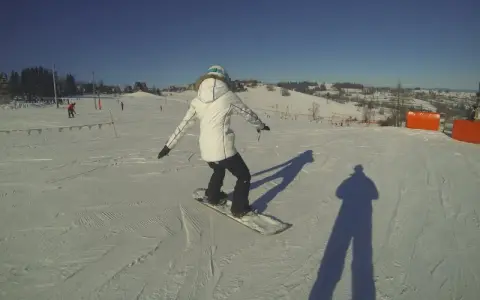 Osoba na snowboardzie na stoku narciarskim, ucząca się w szkole narciarskiej Budzyńskich. Słoneczny dzień, czyste niebo.