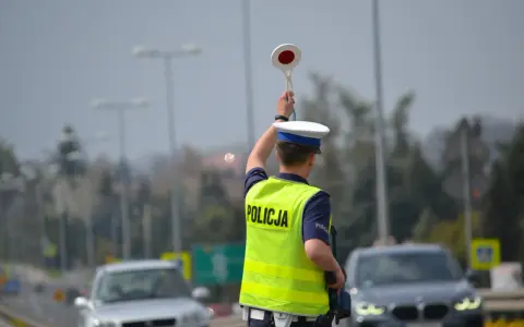 Policjant kieruje ruchem, podnosząc znak stop. W tle widać samochody i słupy oświetleniowe, sugerujące podwyższone **stopnie alarmowe**.