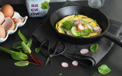 Jak zrobić dobry omlet? Oto inspiracja: puszysty omlet z rzodkiewką, ogórkiem i ziołami na czarnej desce.