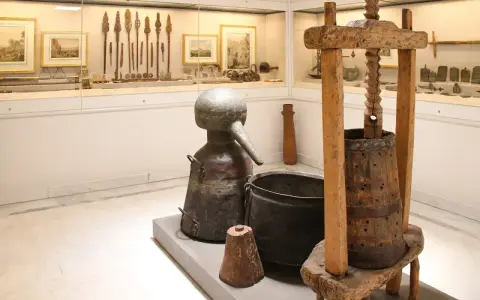 W muzeum Ateny: zabytkowy alembik, kadź i prasa do winogron. W tle ekspozycja narzędzi i obrazów.