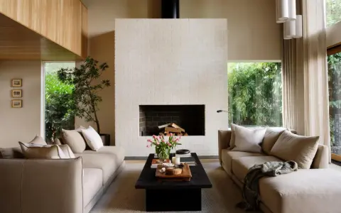 Salón de estilo japandi con sofás beige, chimenea de ladrillo blanco y mucha luz natural.
