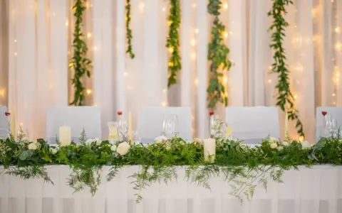 Stół weselny w stylu greenery, udekorowany bujną zielenią, białymi różami i świecami. Tło tworzą zwiewne zasłony i delikatne światełka.