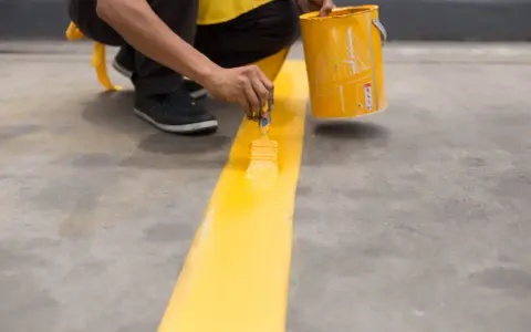 Persona pintando una línea amarilla sobre suelo de cemento sin pulir, usando una brocha y un cubo de pintura.