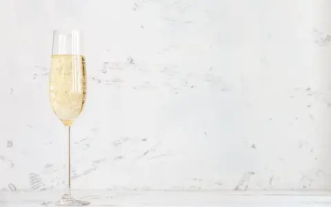 Ett glas champagne med bubblor, kanske en Jaques Selosse, står på ett vitt bord mot en ljus, texturerad bakgrund.