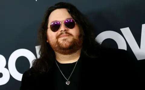 Wolfgang Van Halen w okularach przeciwsłonecznych i z naszyjnikiem w kształcie serca, na tle logo HBO.