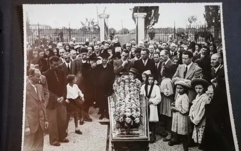 Una folla partecipa a un funerale, con un feretro fiorito al centro. Questa foto post mortem cattura un momento di lutto collettivo.