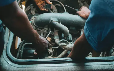 Mechanicy pracują przy silniku, jeden z nich trzyma wtyczkę, drugi dokręca śrubę. Może to być naprawa zaworu par paliwa.