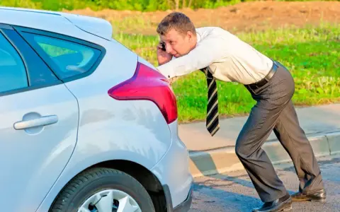 Mężczyzna w garniturze próbuje odpalanie na pych samochodu, trzymając telefon przy uchu.