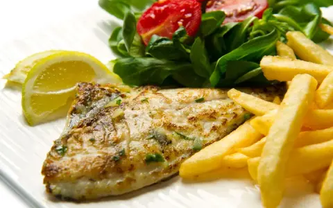 A cooked fish fillet with french fries, salad, and lemon slices on a white plate.