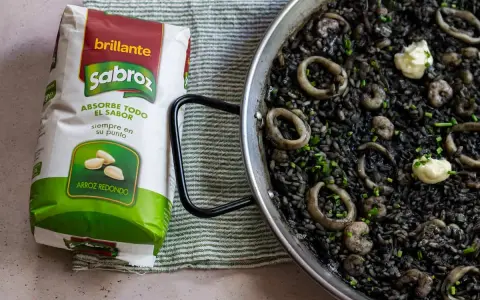 Arroz negro con calamares y gambas en paellera, junto a un paquete de arroz redondo Sabroz.
