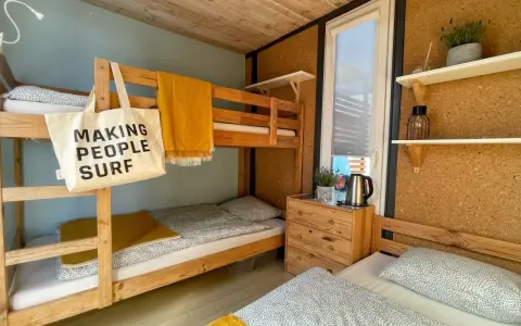 Wnętrze chałupy kempingowej z łóżkiem piętrowym i pojedynczym, żółtym kocem i torbą z napisem "MAKING PEOPLE SURF".