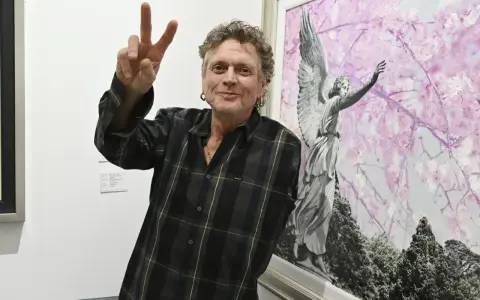 Rick Allen, perkusista Def Leppard, pozuje z uniesionym znakiem pokoju obok obrazu z aniołem i kwitnącymi drzewami.