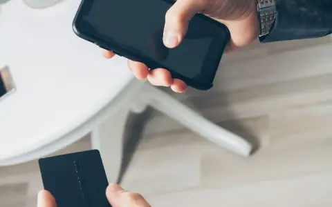 Dłoń trzyma smartfon, druga dłoń czarną kartę płatniczą. Szybkie płatności zbliżeniowe, jak w PKO BP, to wygoda.