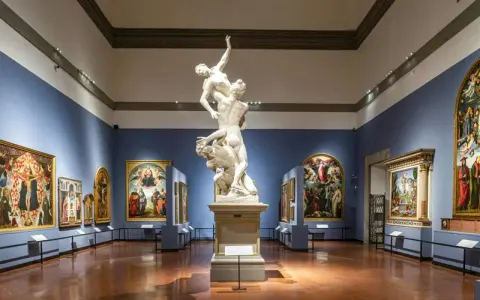 Wnętrze galerii sztuki we Florencji. Marmurowa rzeźba na piedestale, otoczona renesansowymi obrazami na niebieskich ścianach.