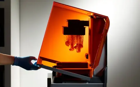 A gloved hand opens the orange lid of a stereolithography 3D printing machine, revealing intricate parts being built layer by layer.