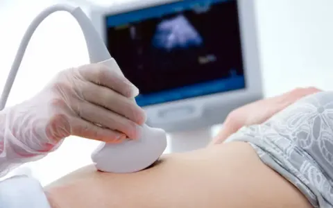 Ręka w rękawiczce trzyma sondę USG, która bada brzuch. Na ekranie widać obraz z badania, być może to test pappa.