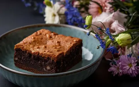 Un délicieux brownie healthy, parsemé de noix, dans un bol bleu, avec un bouquet de fleurs colorées en arrière-plan.