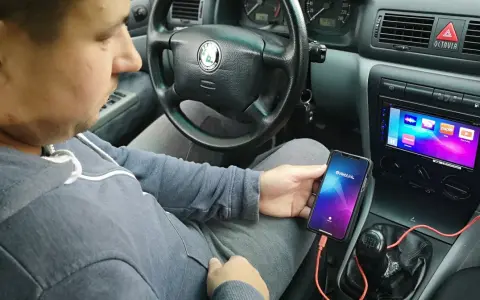 Mężczyzna w samochodzie pokazuje, jak połączyć telefon z autem za pomocą kabla i aplikacji Weblink. Na ekranie radia widoczne są ikony aplikacji.