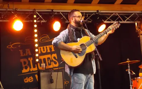 Un homme joue de la guitare sur scène, éclairé par des projecteurs. Le panneau derrière lui annonce "La Nuit du Blues", évoquant la naissance du blues.