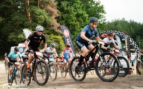 Kolarze biorą udział w wyścigu **zawody MTB**. Widać ich w akcji, jadących po piaszczystej trasie, z lasem w tle.