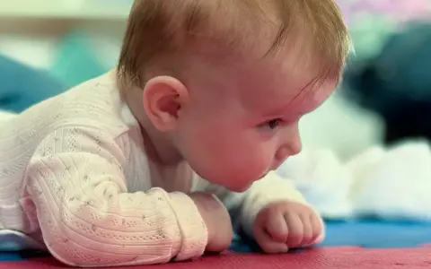 Malutkie dziecko leży na brzuchu, z zaciekawieniem spoglądając przed siebie. Jego duża głowa u dziecka jest lekko pochylona, a drobne rączki podparte o podłoże.