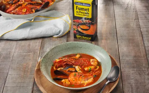 Un plato humeante de zarzuela de pescado, con gambas, mejillones y calamares, junto a un cartón de fumet de pescado.