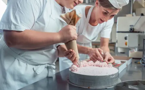 Deux pâtissières décorent un gâteau rose lors d'un cours de pâtisserie en ligne. L'une utilise une poche à douille, l'autre des fruits.