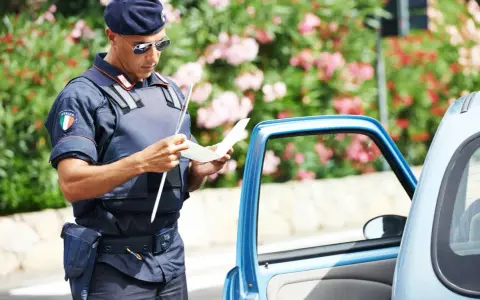 Agente di polizia italiana con giubbotto antiproiettile e berretto blu, controlla un verbale vicino a un'auto blu. Potrebbe essere un controllo legato all'art. 180 cds.